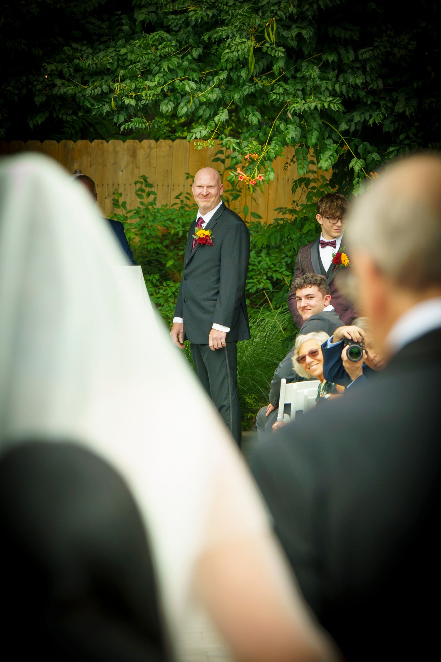 Groom in dark suit smiling at bride walking down the aisle at outdoor wedding ceremony with guests watching.