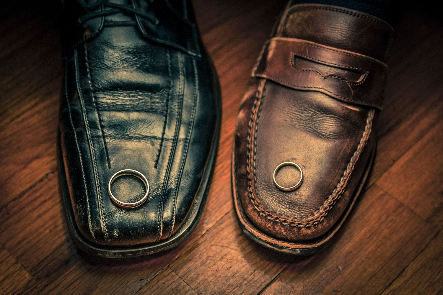 Two wedding rings resting on men's dress shoes, one black oxford and one brown loafer, on a wooden floor.