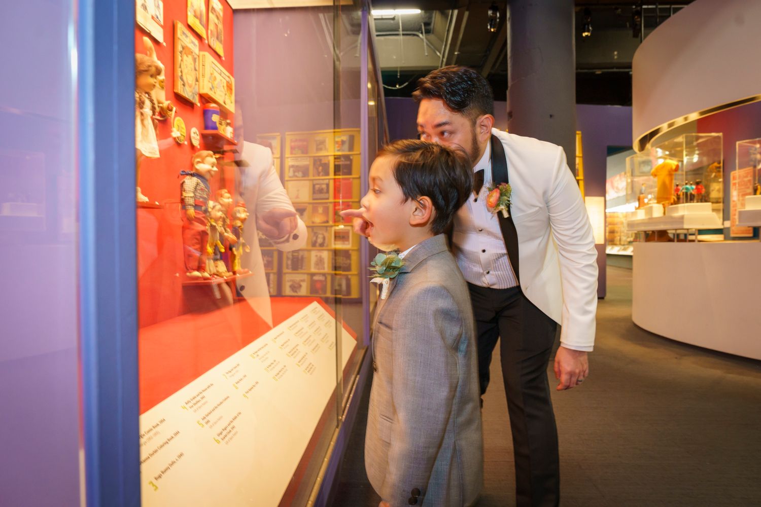 Groom in white tuxedo showing young boy exhibits at museum display case during wedding day.