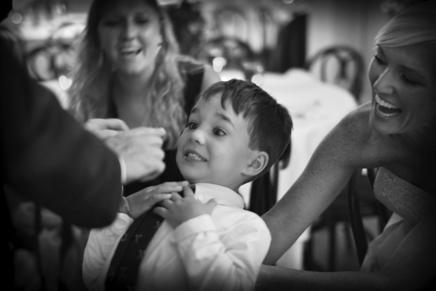 Young boy in dress shirt laughing with bridesmaids at wedding reception, black and white.