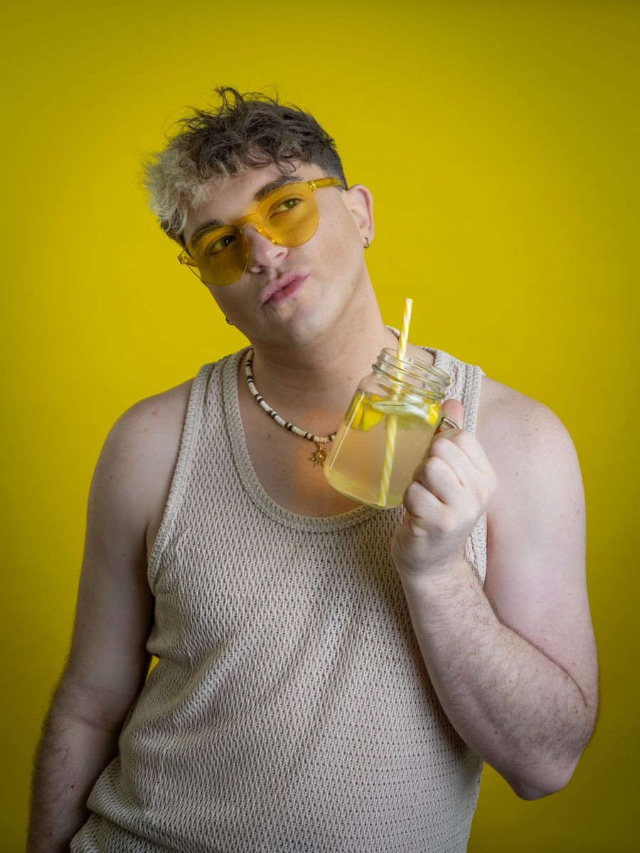 Person in beige mesh tank top with yellow sunglasses holding mason jar drink with straw on yellow background.