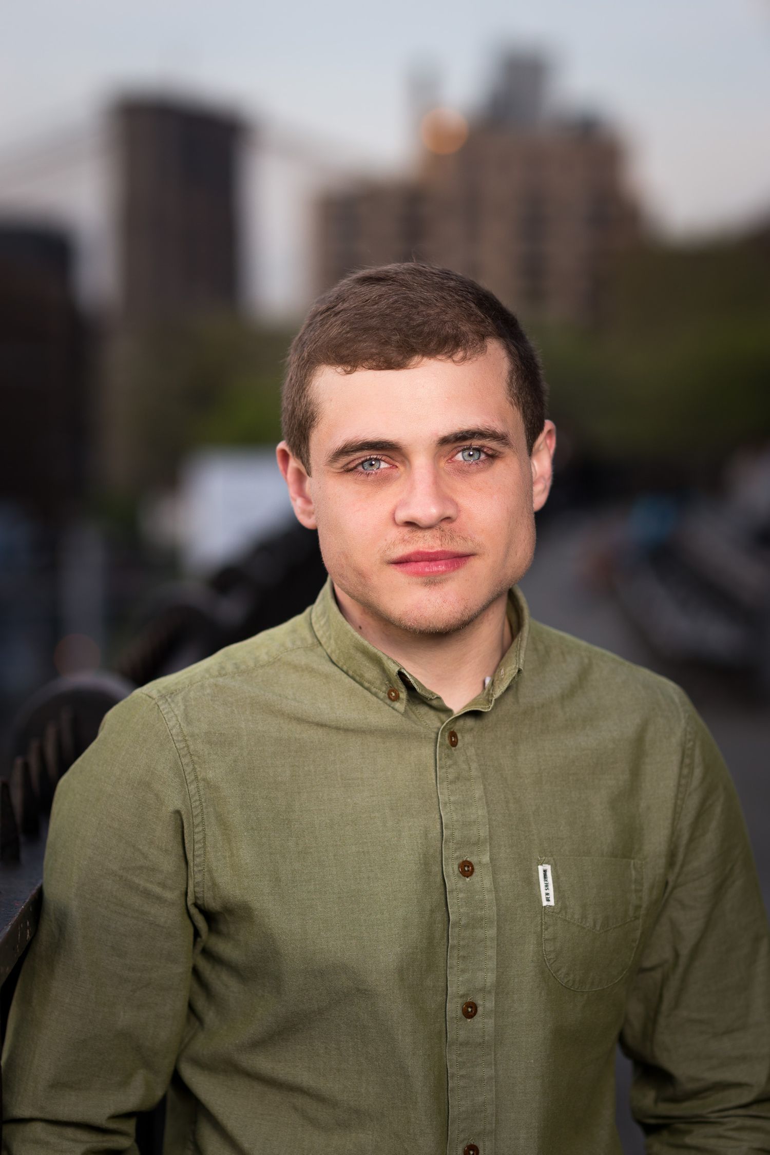 Young man in olive green shirt posing outdoors with urban skyline background.