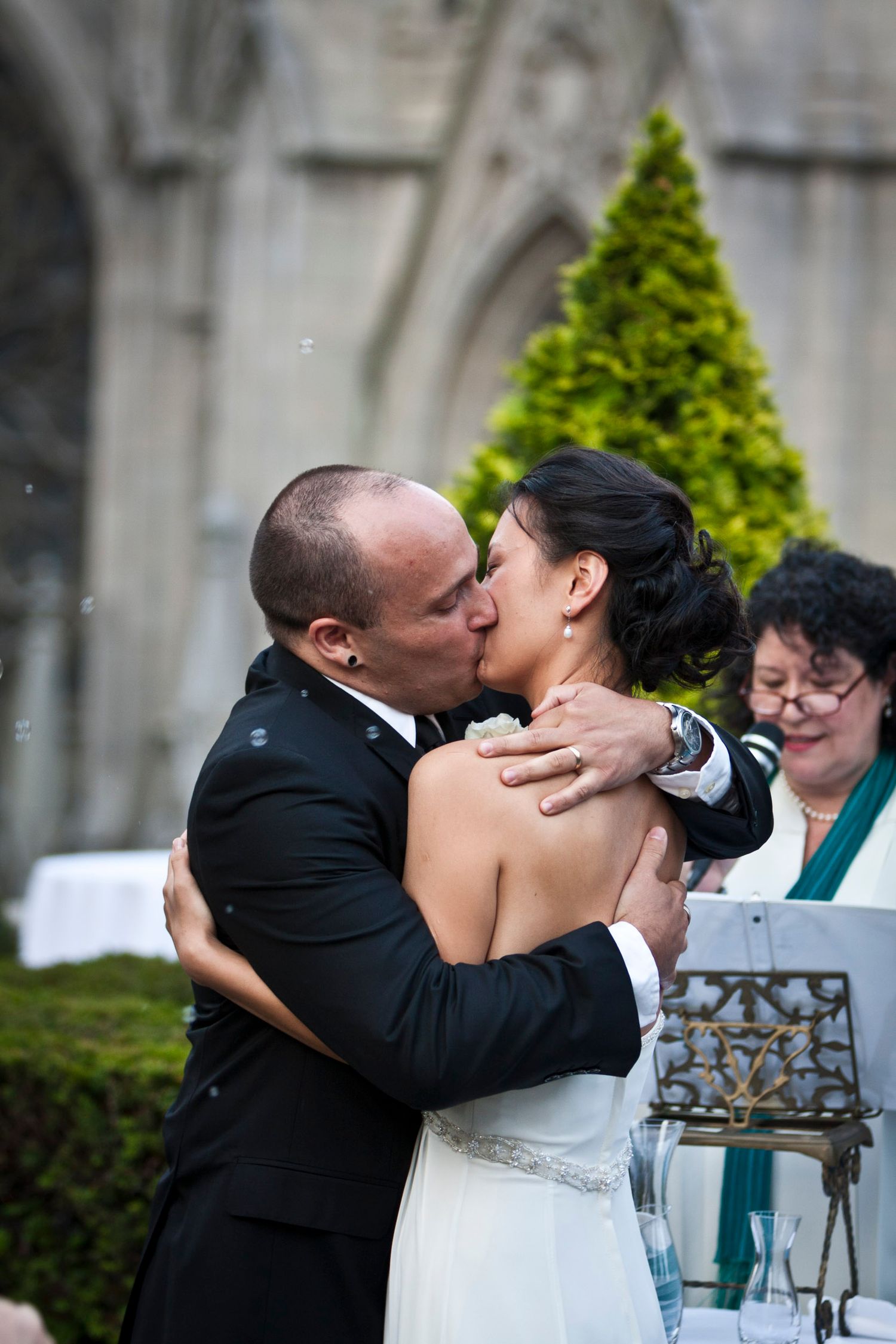 Groom in black suit shares first kiss with bride in strapless gown at outdoor wedding ceremony.