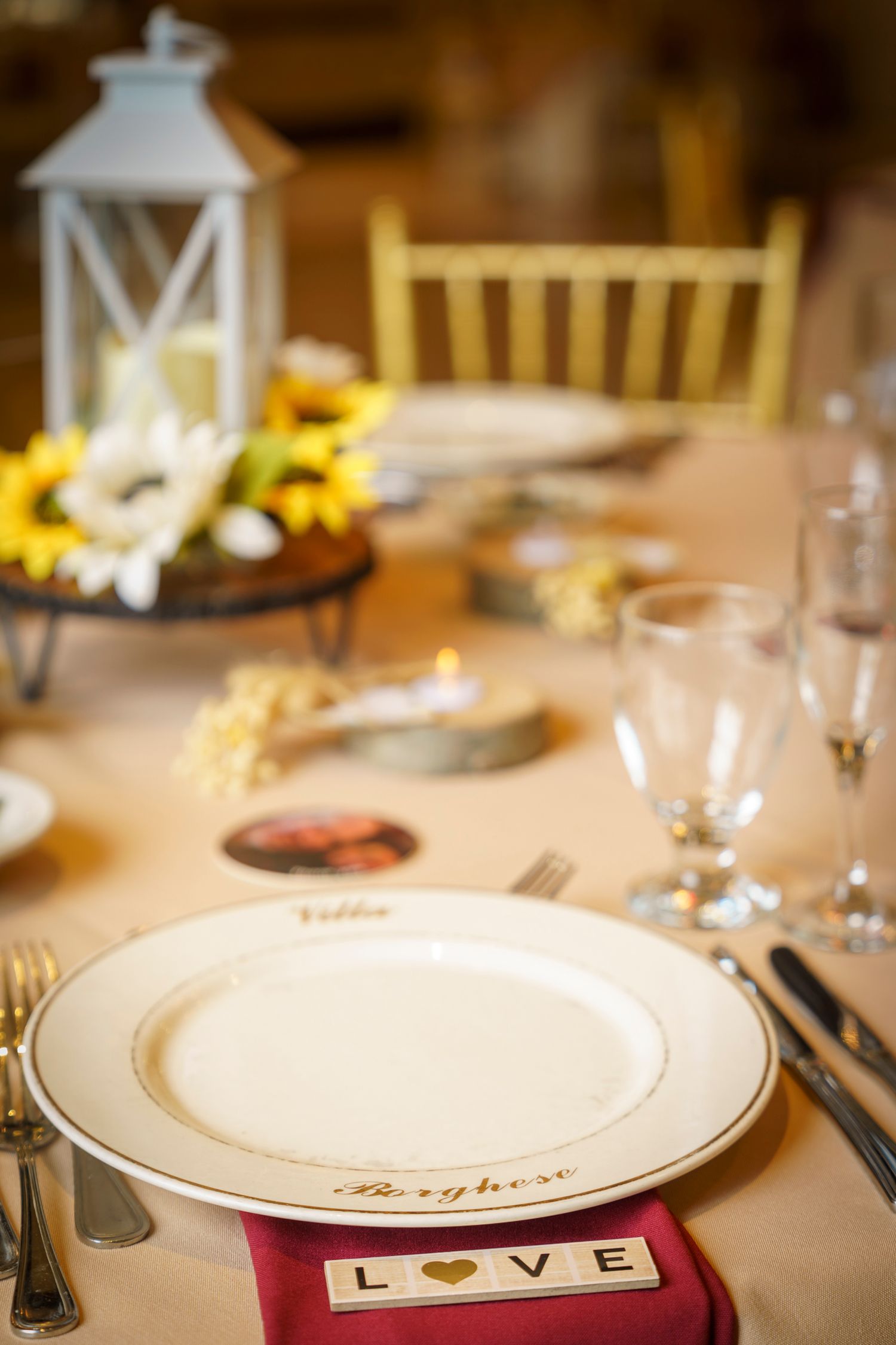 Elegant wedding table setting with a white plate, LOVE tiles, sunflower centerpiece, and white lantern on a cream tablecloth.