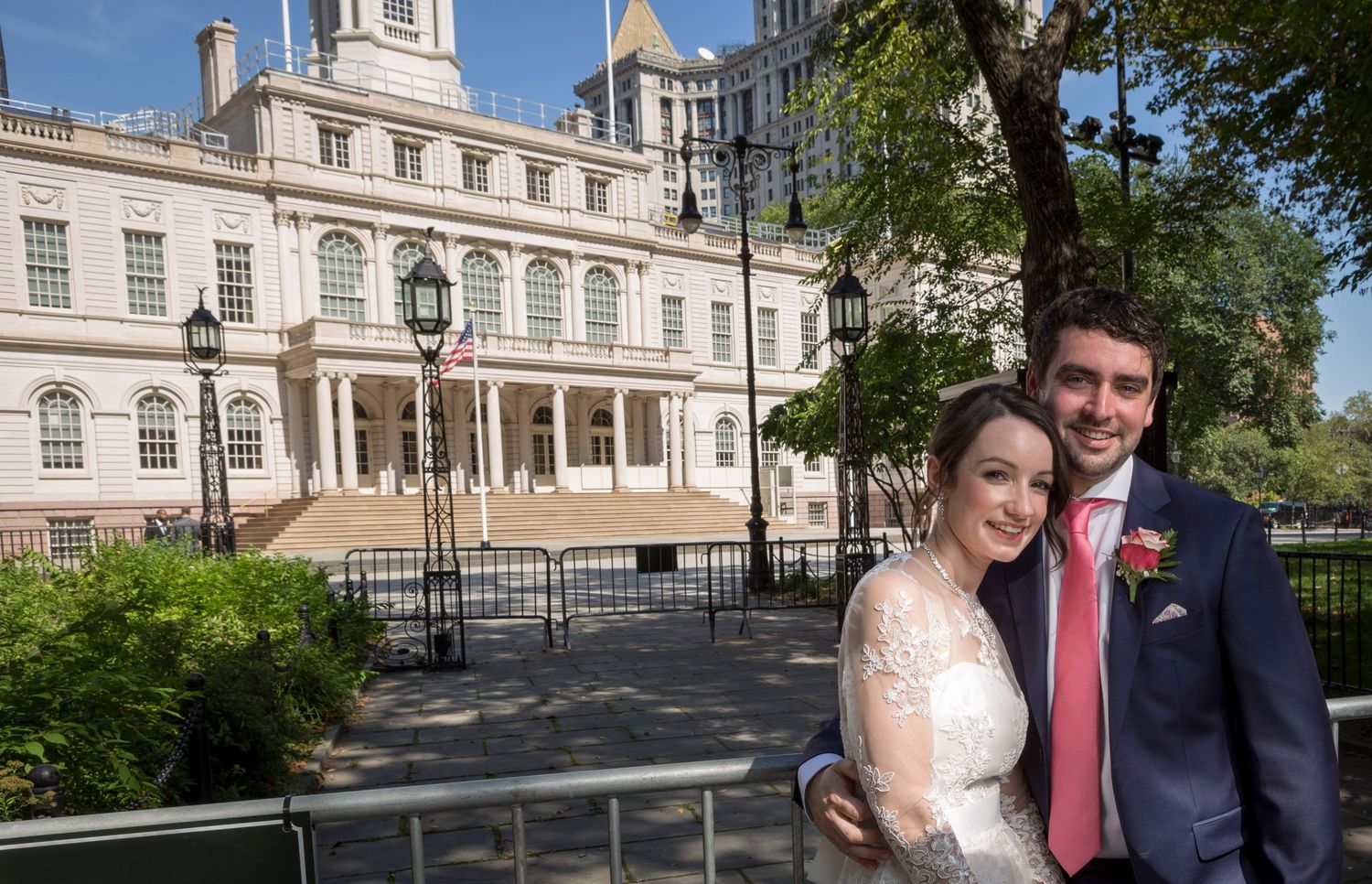 Newlywed couple posing in front of New York City Hall on their wedding day, bride in lace gown and groom in suit.