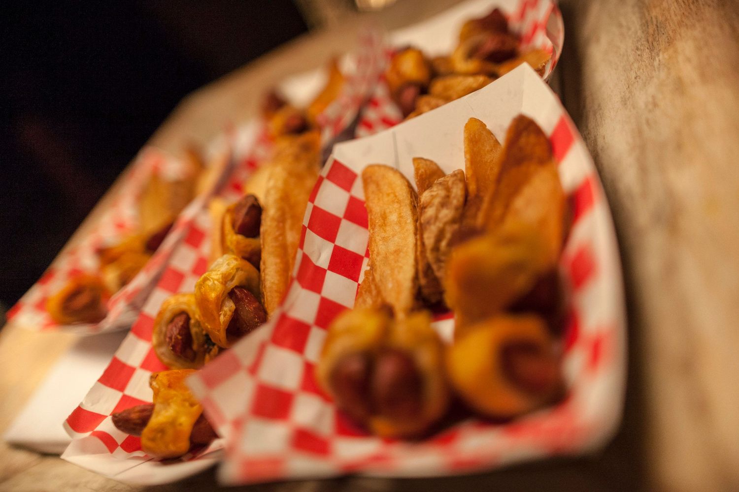 Assorted fried snacks and hot dogs served in red checkered paper-lined baskets at a casual food event.