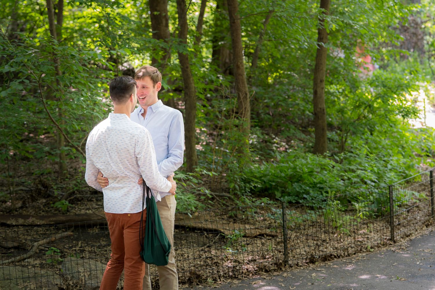 Two men standing together in a lush green park, sharing a tender moment near a black iron fence.