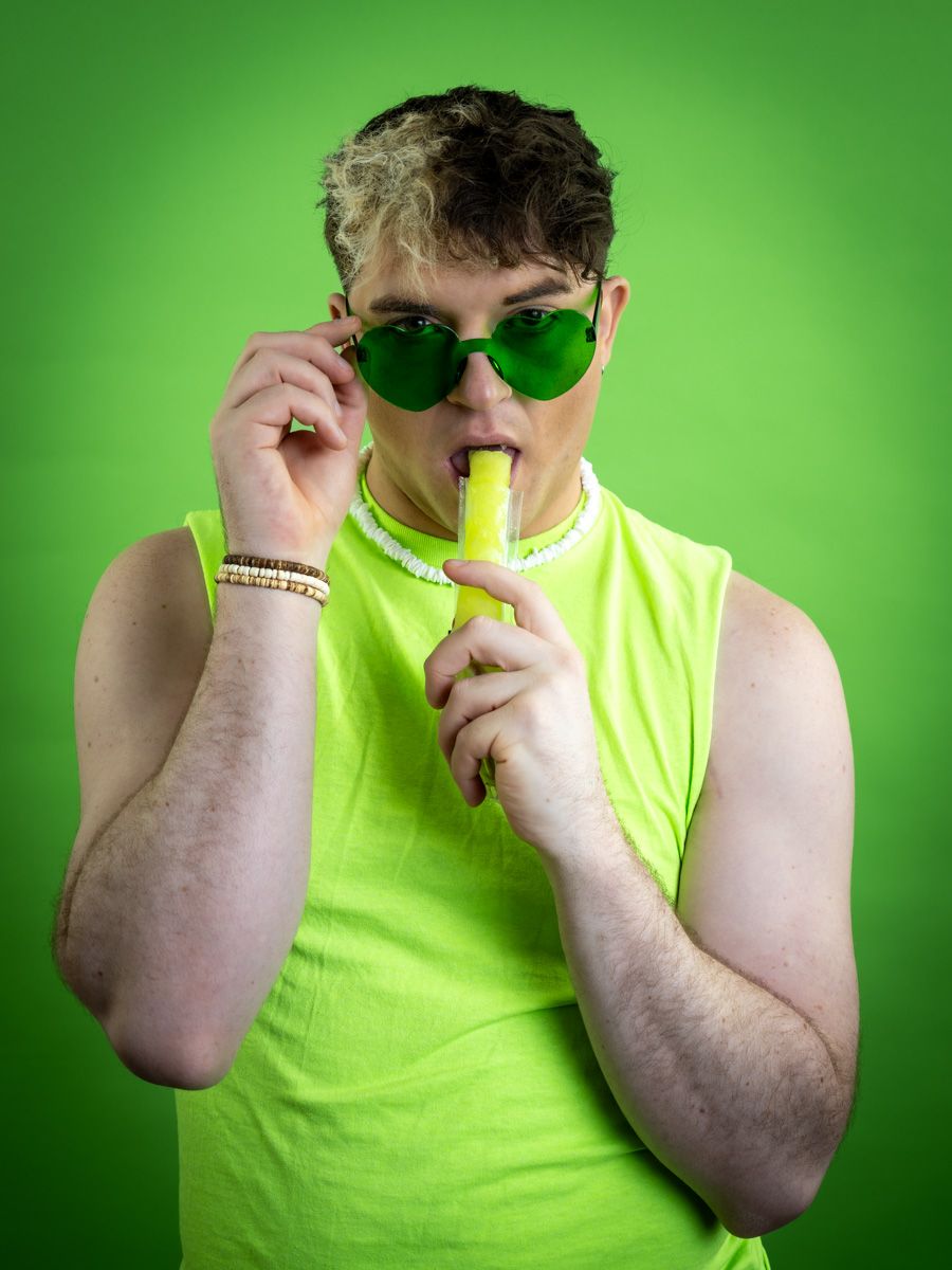 Young man in neon green tank top and heart-shaped sunglasses blowing a party horn against green background.