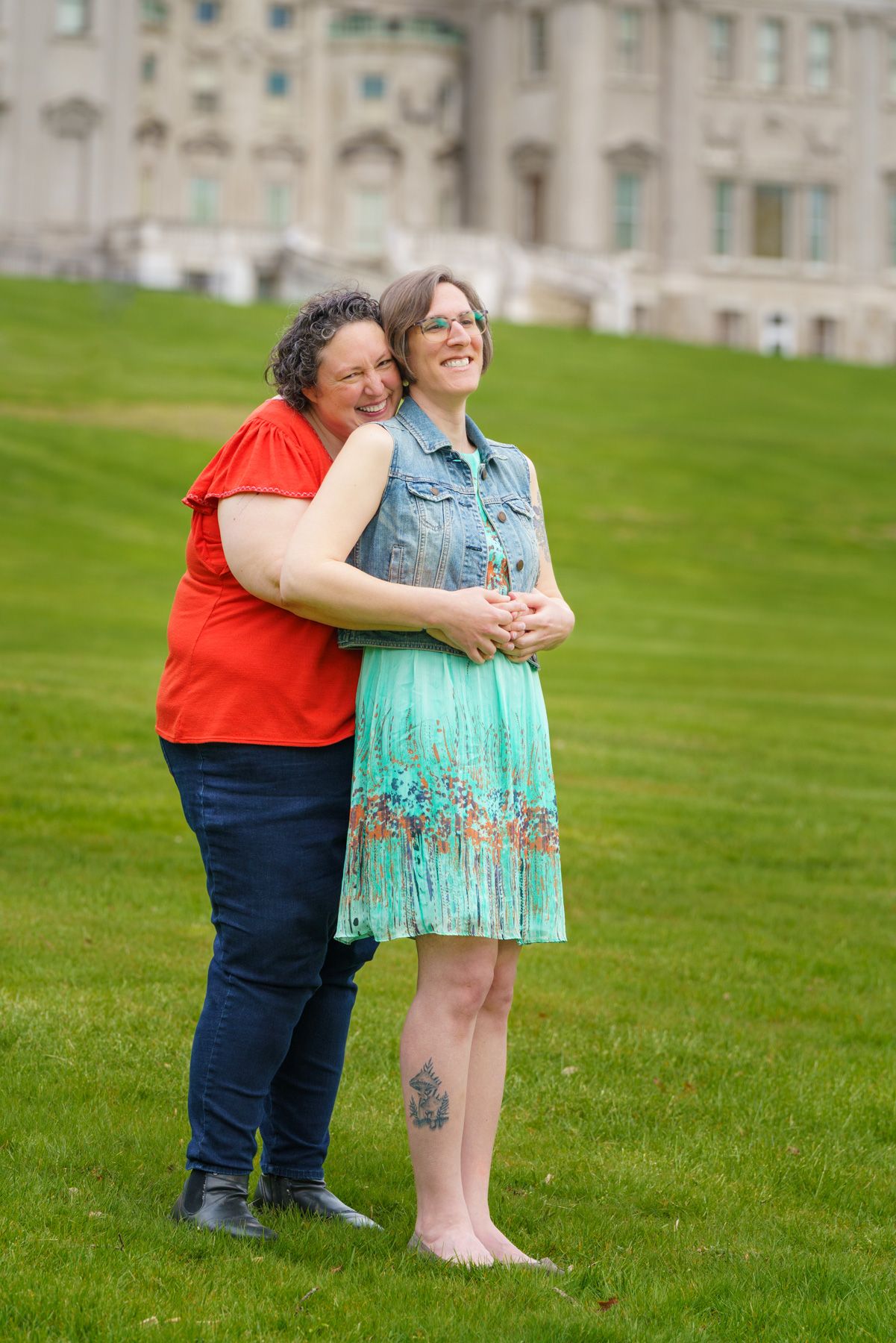 Two women embrace and smile on a green lawn in front of a large stone building on a sunny day.