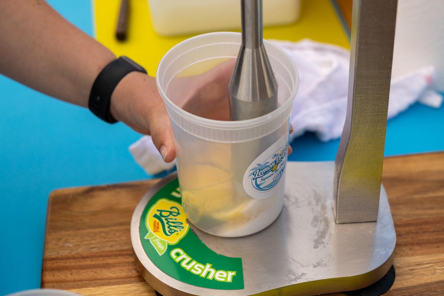 Hand using immersion blender in plastic cup on a Crusher machine base on wooden cutting board.