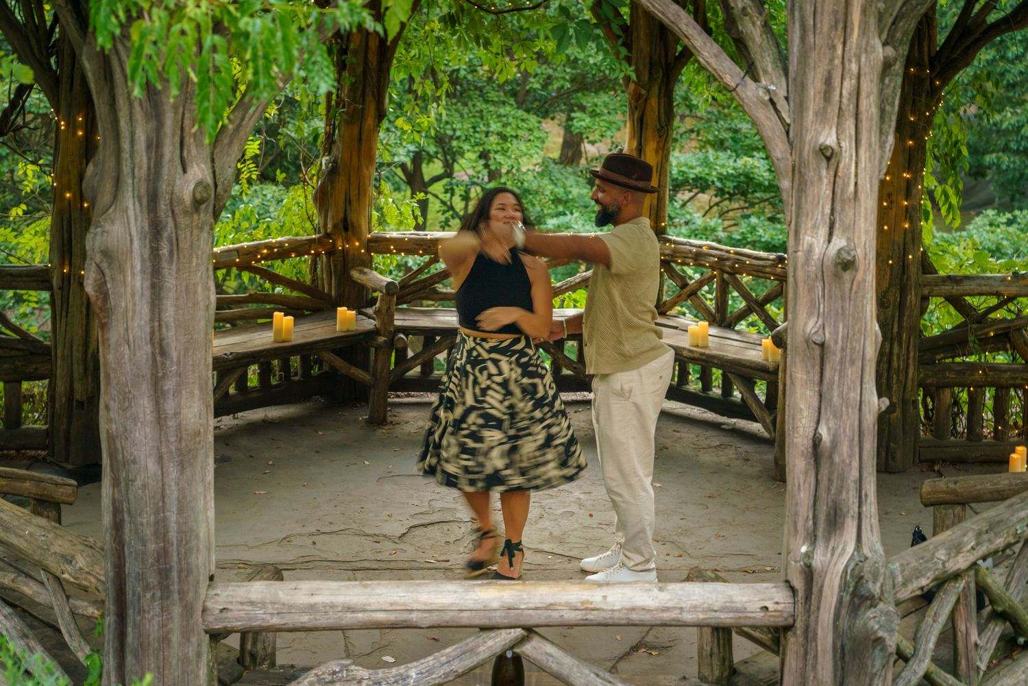 Couple dancing together on a wooden gazebo surrounded by lush green trees in a scenic outdoor setting.