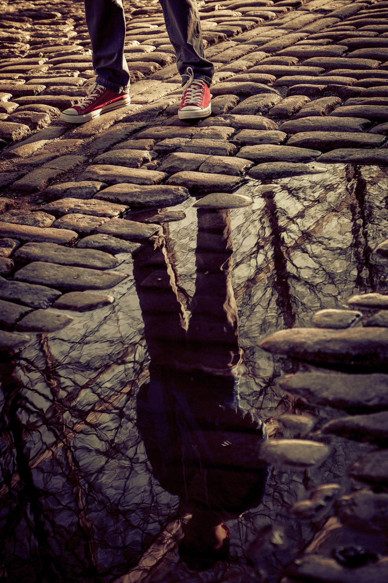 Artistic reflection of a person in red Converse sneakers captured in a cobblestone puddle.