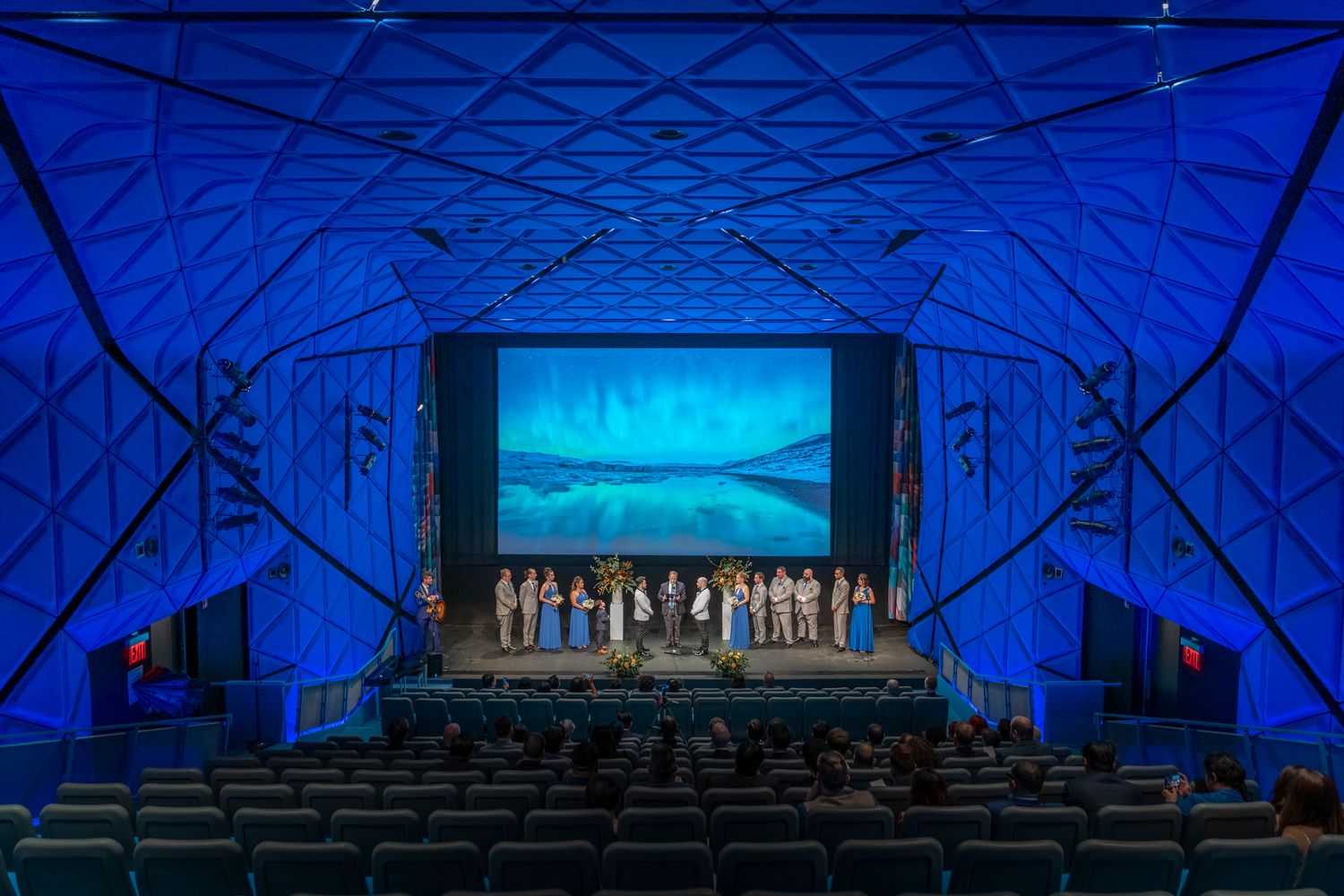 Large blue-lit auditorium with audience watching a group on stage with a scenic backdrop.