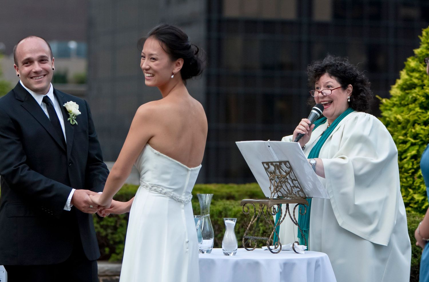 Bride and groom hold hands while female officiant reads vows at outdoor wedding ceremony.