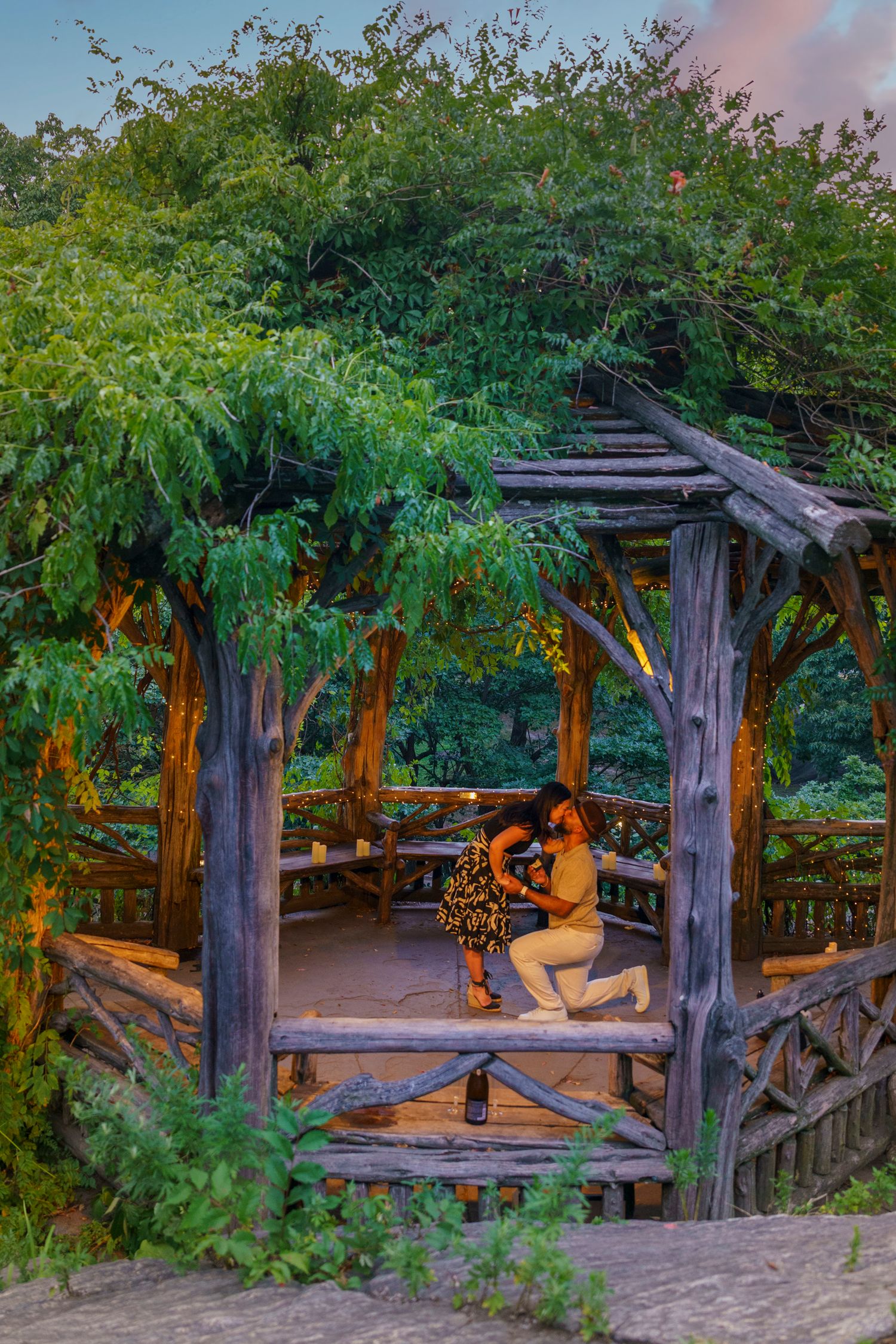 A couple shares a romantic proposal moment in a rustic wooden gazebo surrounded by lush green trees at dusk.