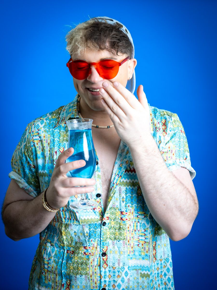 Man in red heart sunglasses and tropical shirt holds blue drink, covering mouth with hand against blue background.