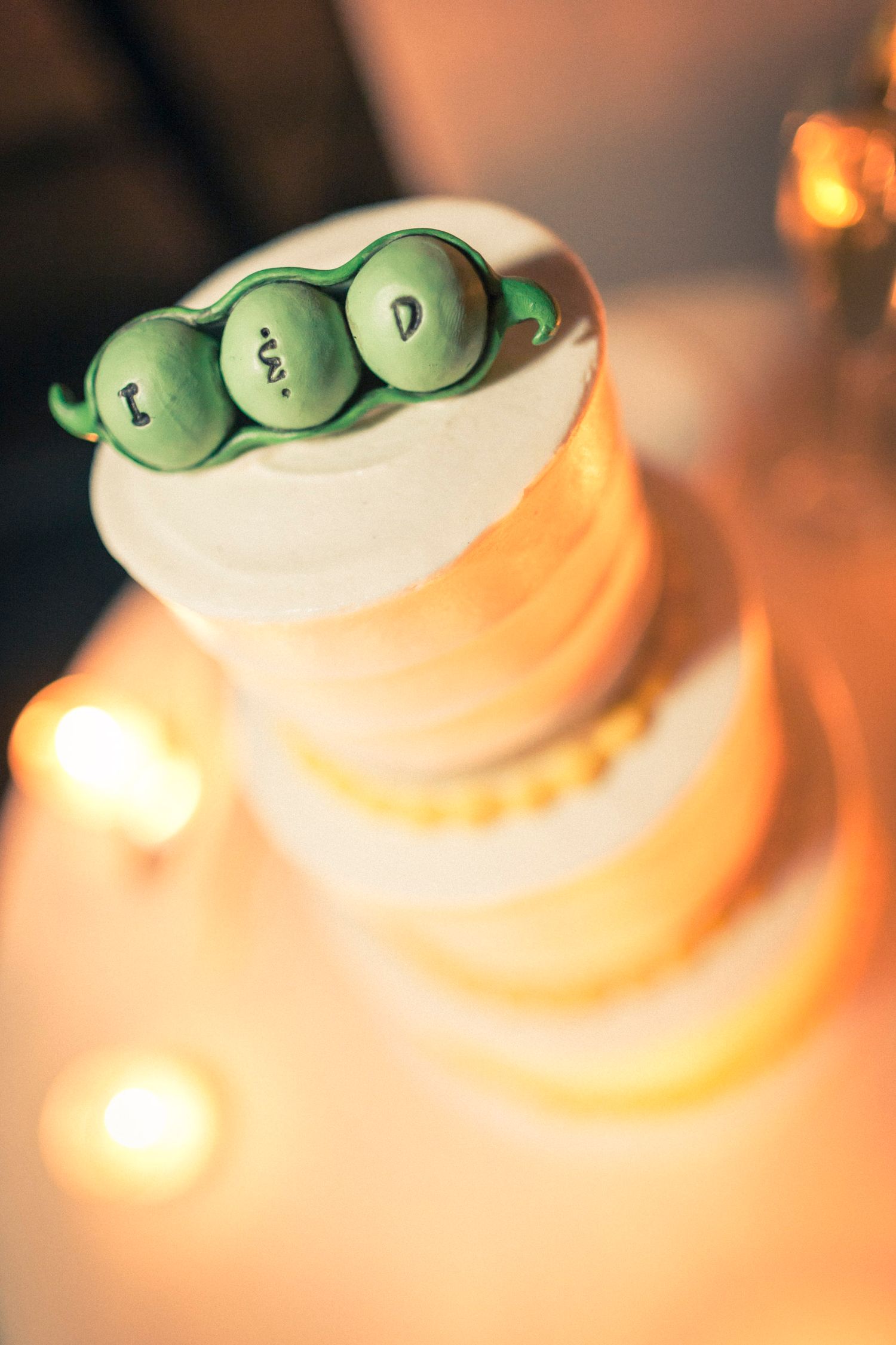 Three-tier wedding cake topped with fondant peas in a pod bearing initials, surrounded by warm candlelight.