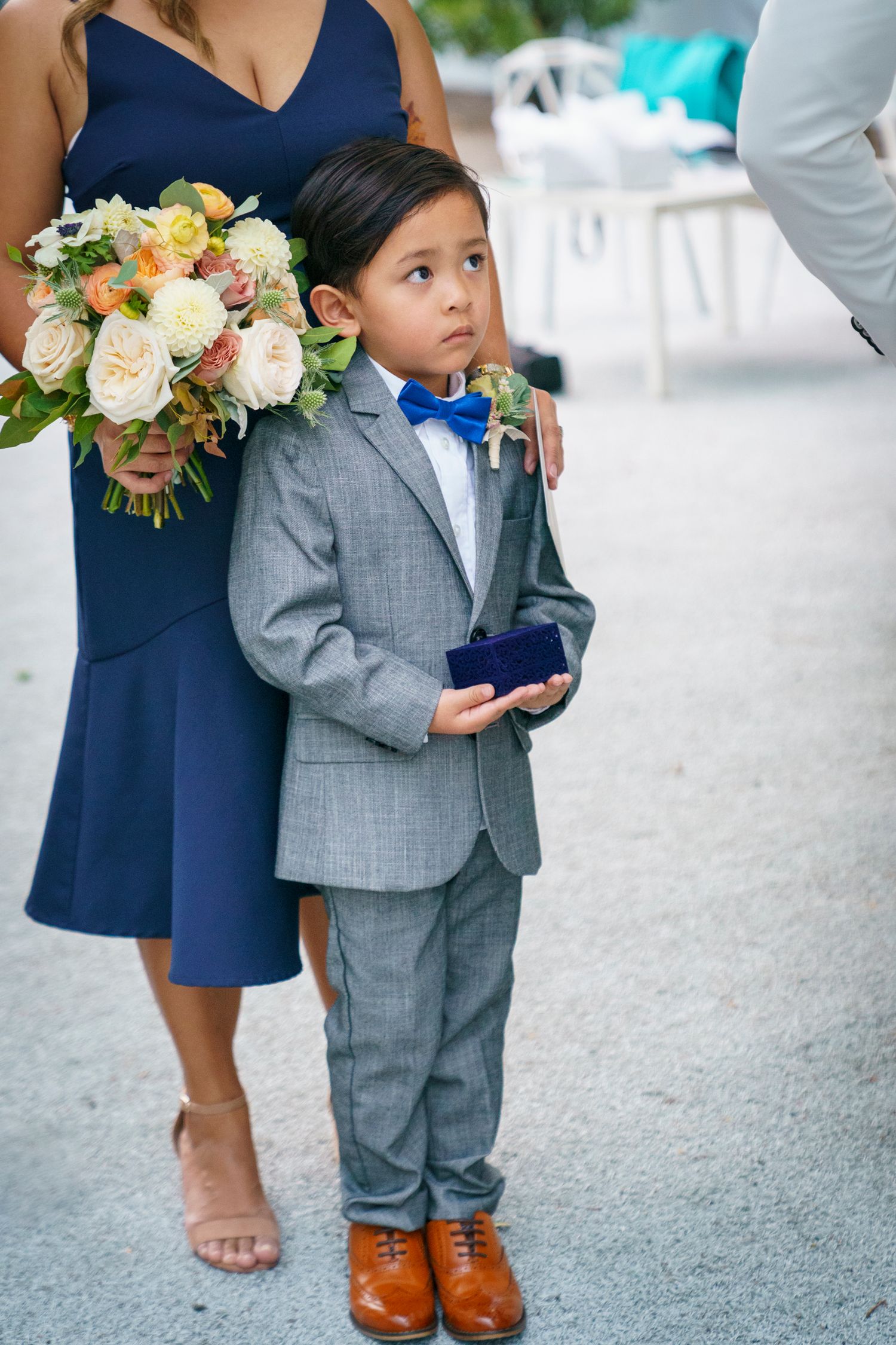 Adorable ring bearer in gray suit and blue bow tie holds velvet ring box at an outdoor wedding ceremony.