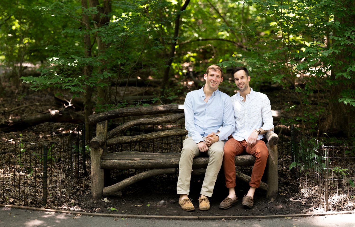 Two men smiling and sitting together on a rustic wooden bench surrounded by lush green trees outdoors.
