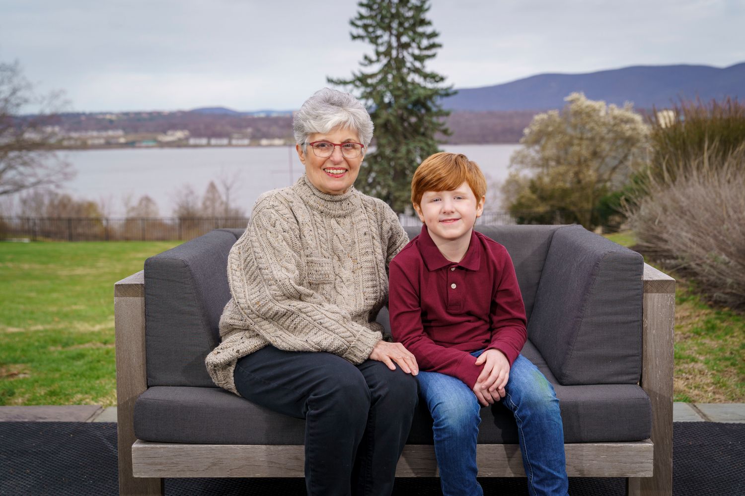 Grandmother and red-haired grandson smiling on outdoor sofa with lake and mountains in background.