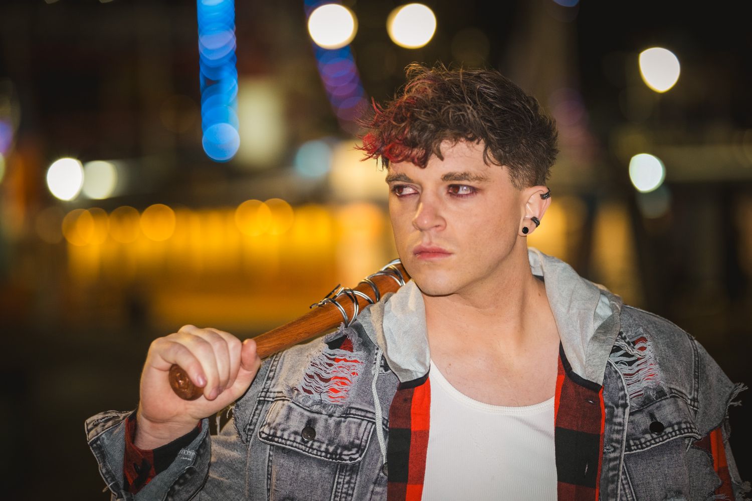 Young man with curly dark hair wearing a distressed denim jacket over a plaid shirt holds a cigar against a blurred city lights background.