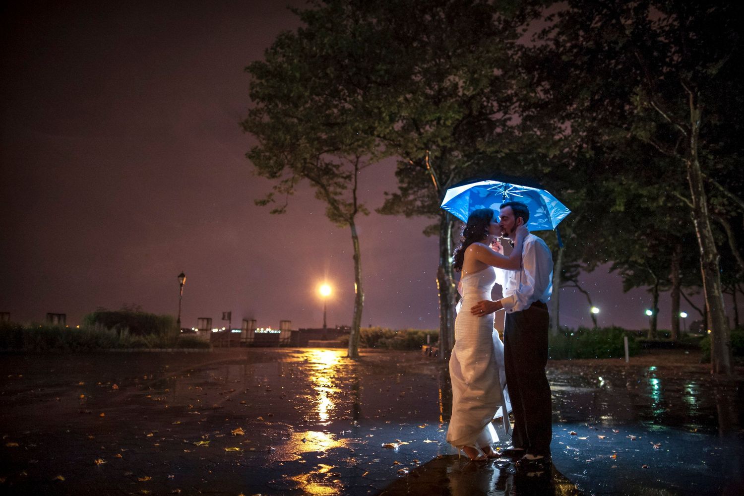 Bride and groom share a romantic kiss under a glowing blue umbrella in the rain at night.