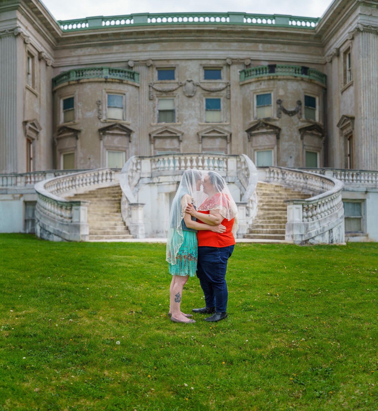 Couple sharing a kiss under a veil on green lawn before a grand neoclassical stone building with curved staircases.