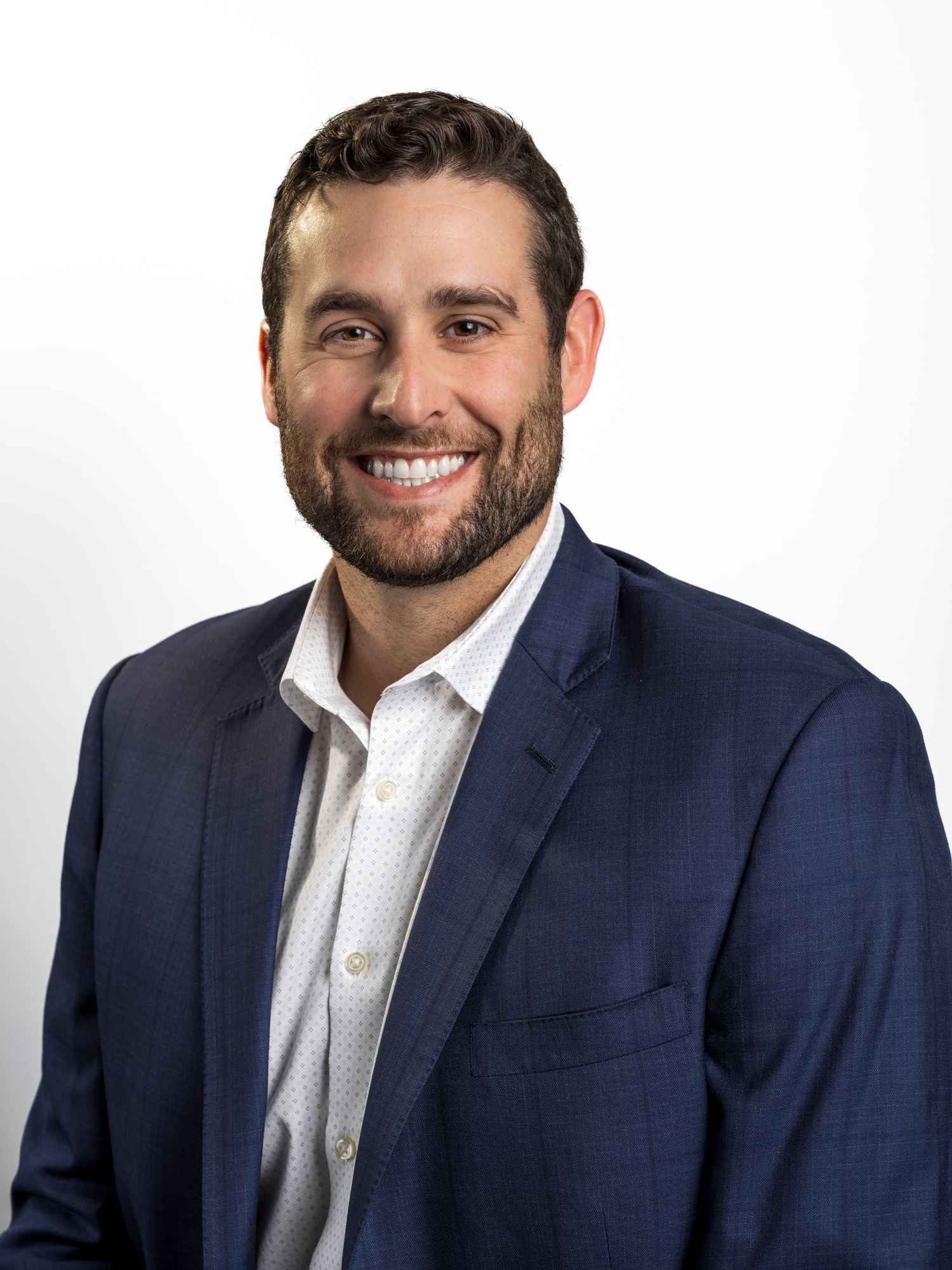 Professional headshot of a smiling man with beard wearing a navy blue blazer and white dress shirt against a light background.