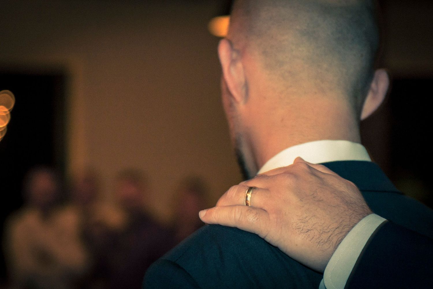 A man in a suit seen from behind with a hand resting on his shoulder wearing a gold wedding ring.