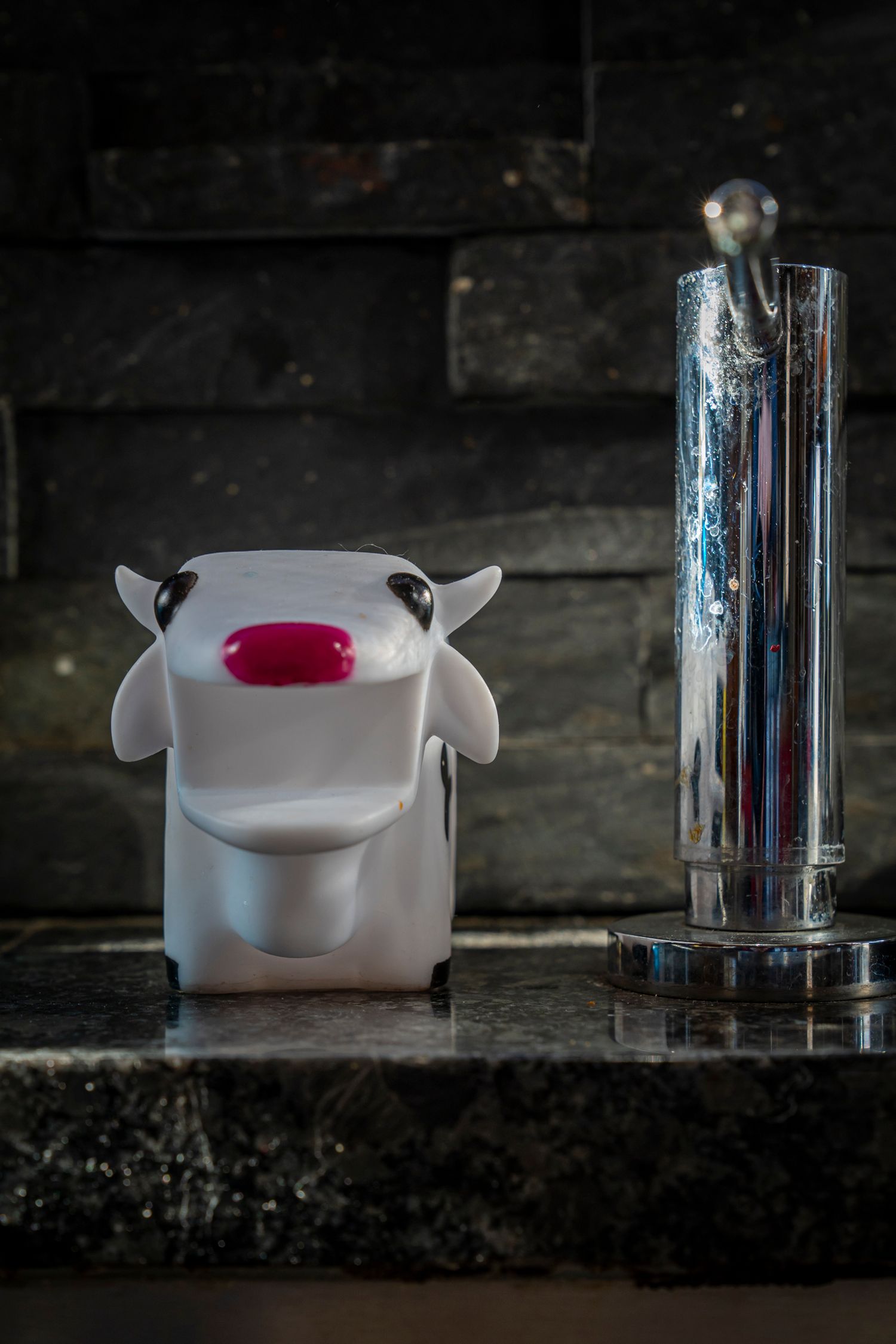 White cow toy figurine with pink nose beside a chrome beer tap on dark stone surface.