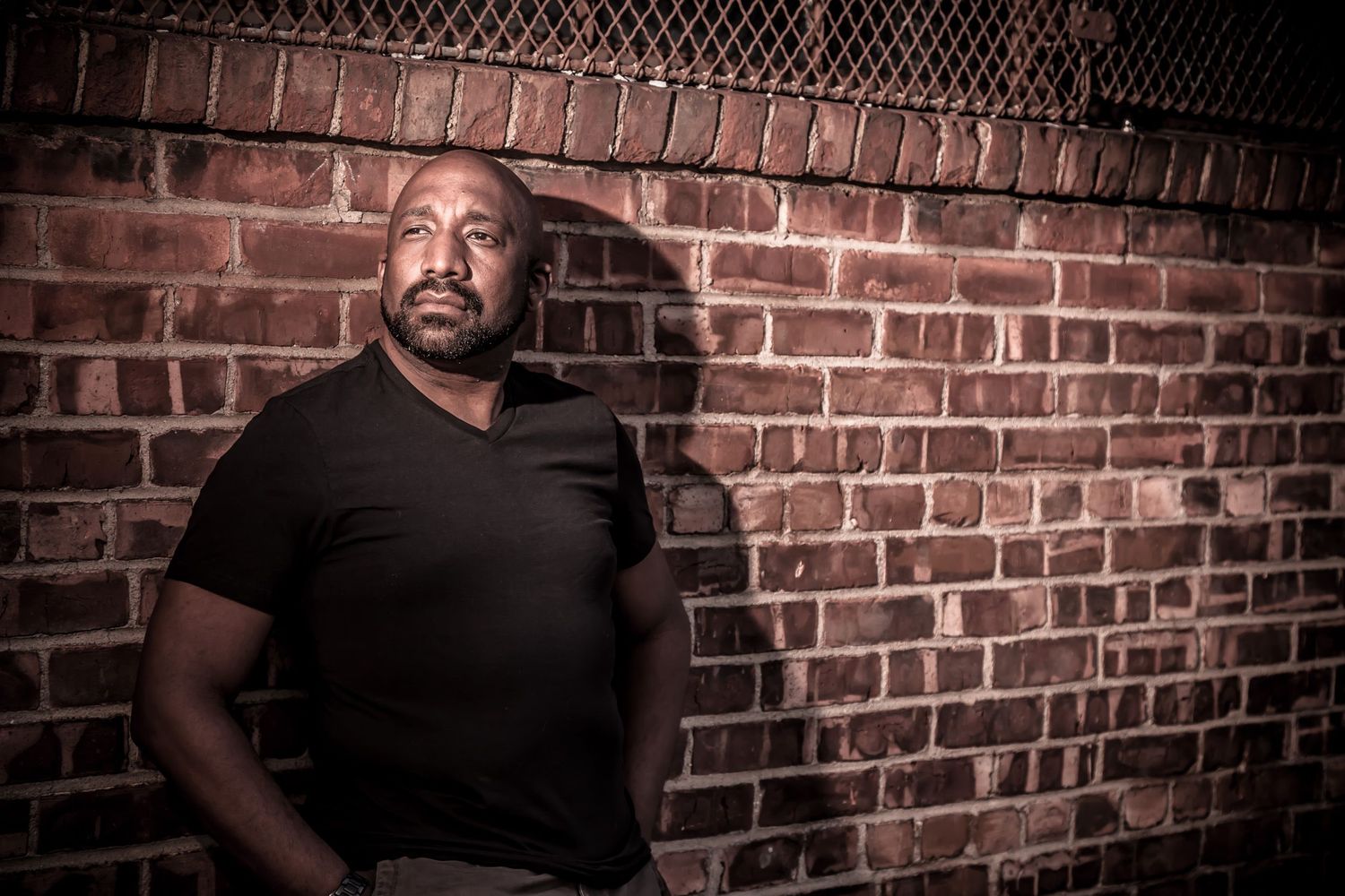 African American man in black t-shirt leaning against brick wall at night.