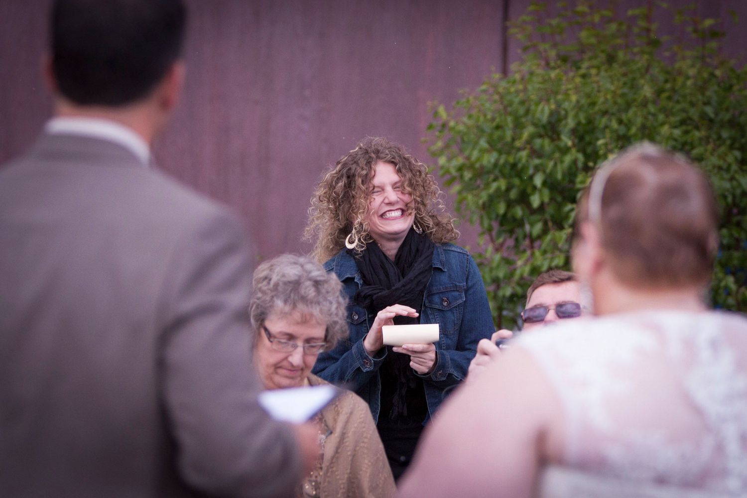 Guests laughing and celebrating at an outdoor wedding ceremony, with the bride visible in the foreground.