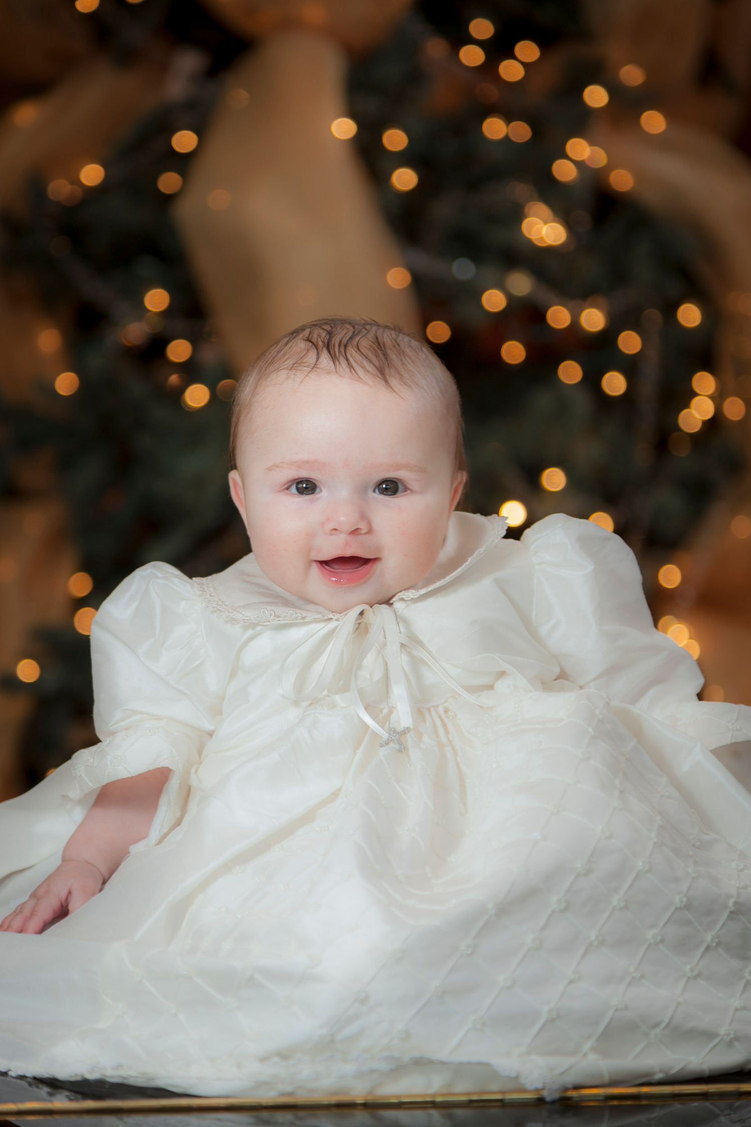 A happy baby in a white christening gown sits before a glowing Christmas tree with bokeh lights.