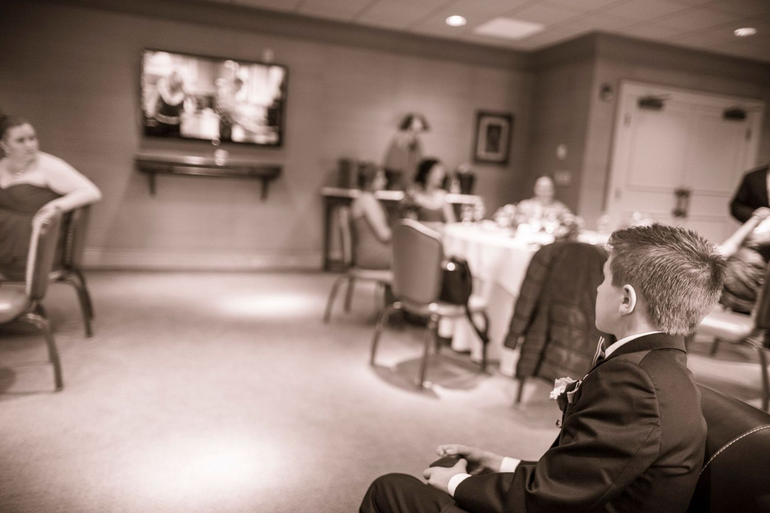 Man in suit seated at wedding reception watching TV screen in elegant banquet hall.