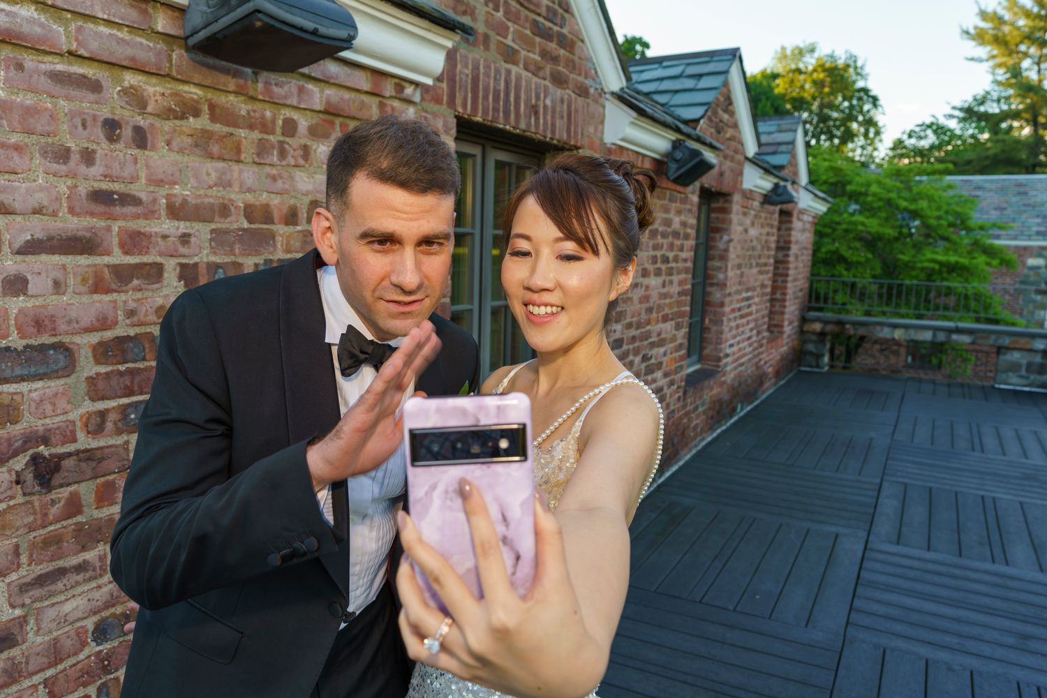 Couple in formal attire taking a selfie with a pink smartphone outside a brick building.