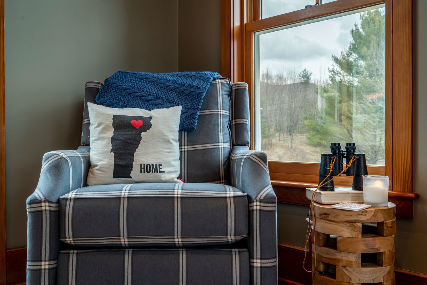Cozy plaid armchair with a Home pillow and blue throw blanket beside a window with binoculars on a wooden stool.