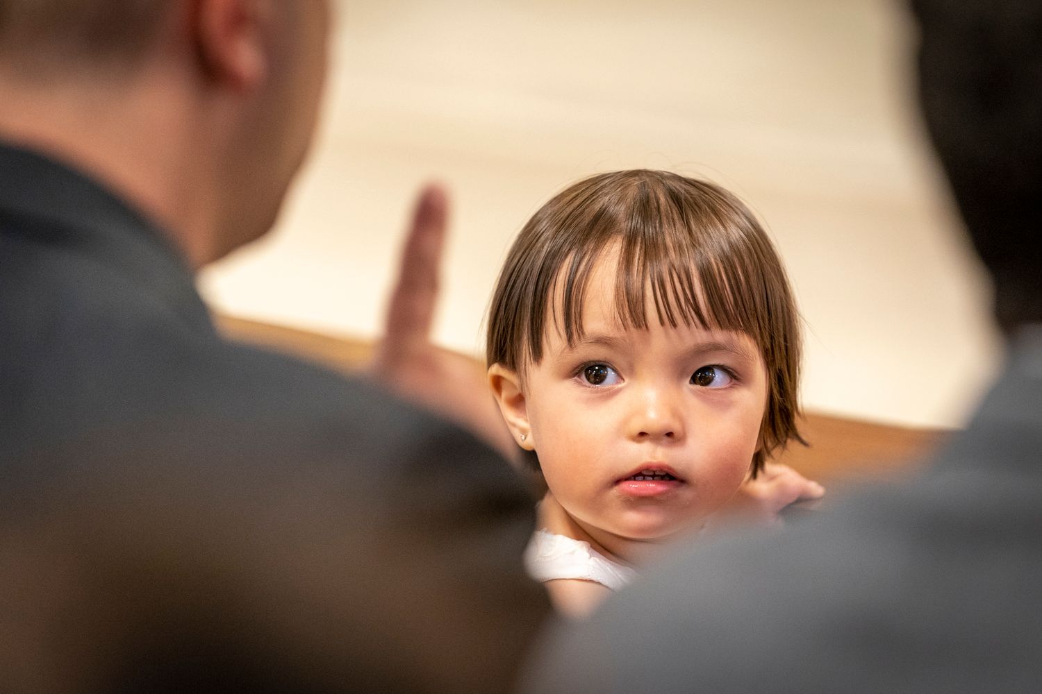 A young toddler with brown hair looks up attentively at an adult gesturing with their hand indoors.