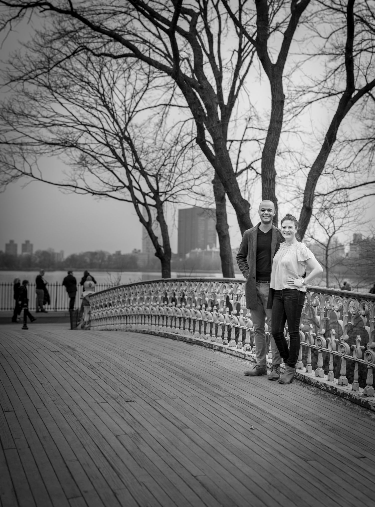 Black and white photo of a couple posing on a wooden bridge in Central Park, NYC, with bare trees and skyline backdrop.