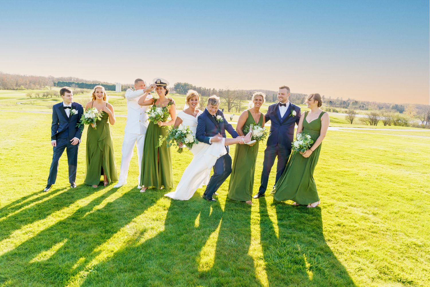 Wedding party in green gowns and navy suits celebrating on lush lawn with scenic countryside backdrop on sunny day.