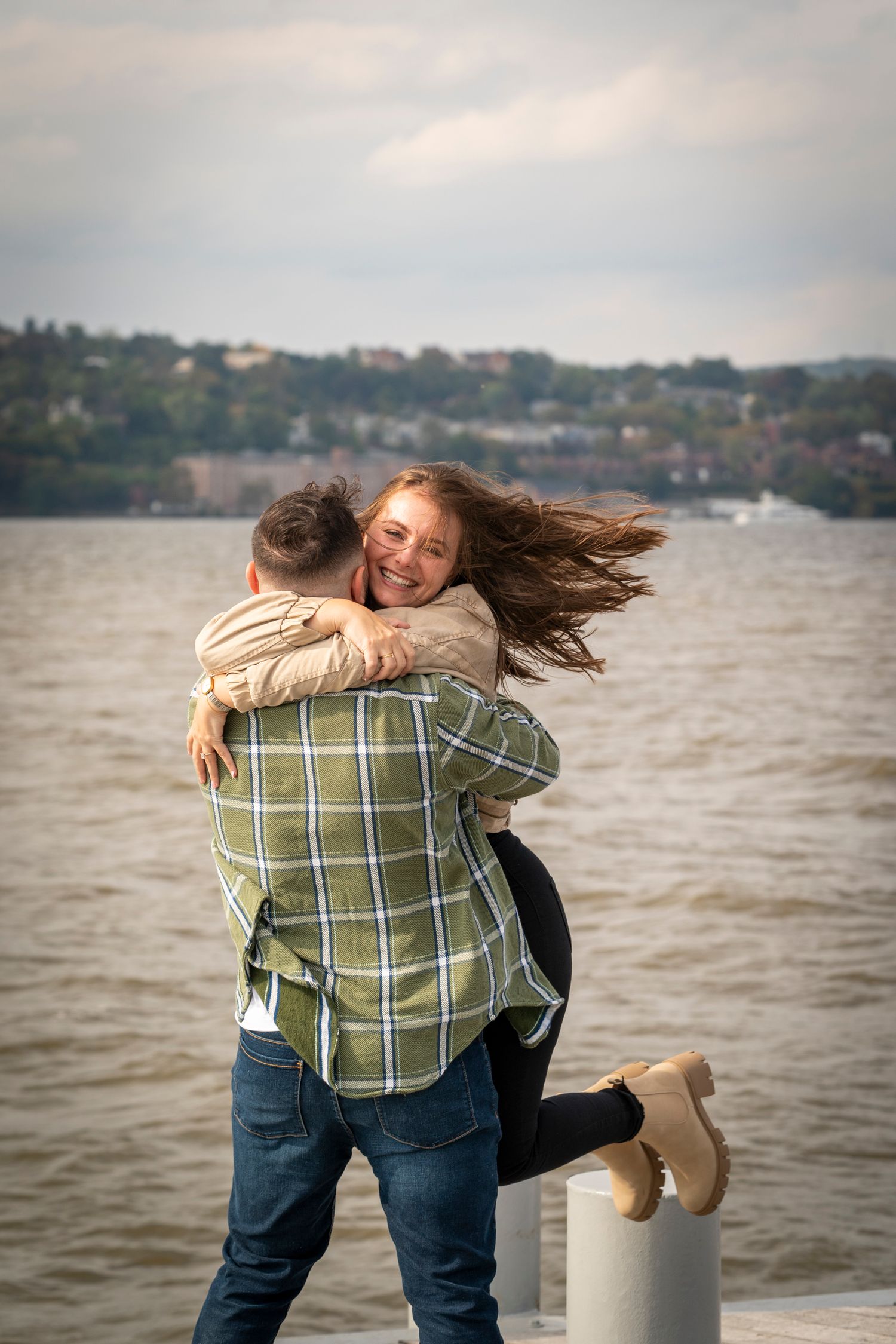 Happy couple embracing on a waterfront pier, woman smiling with hair blowing in the wind on a cloudy day.