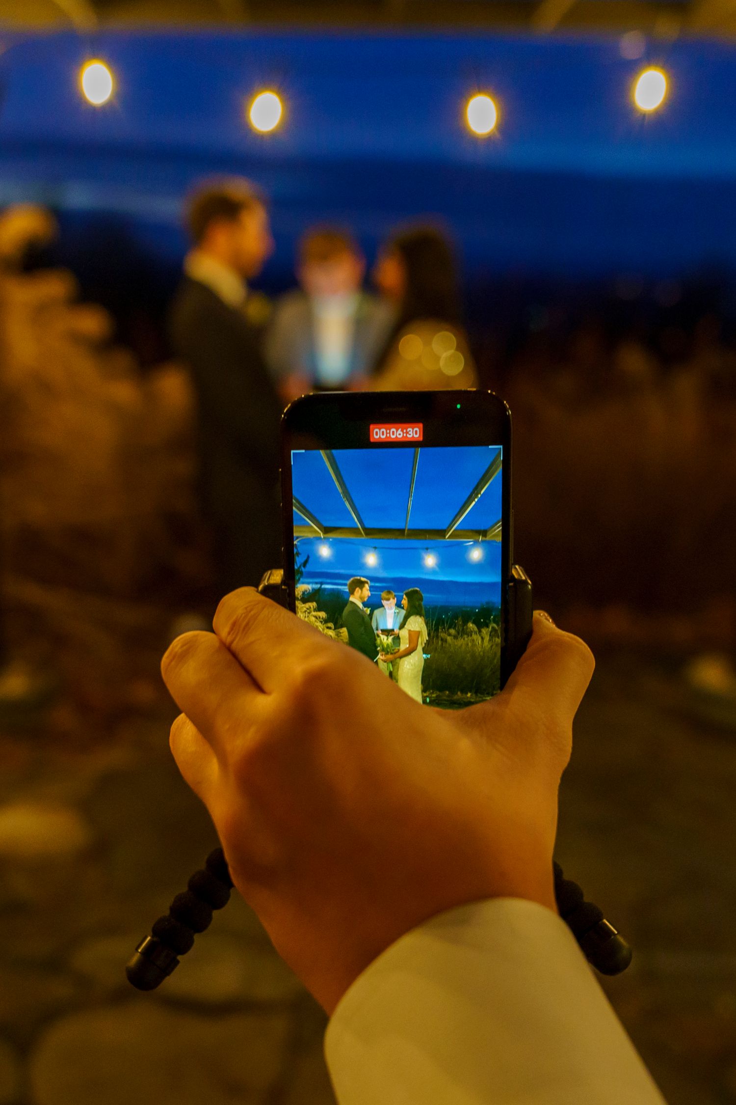 A hand holds a smartphone recording a live wedding ceremony outdoors at night with string lights glowing in the background.