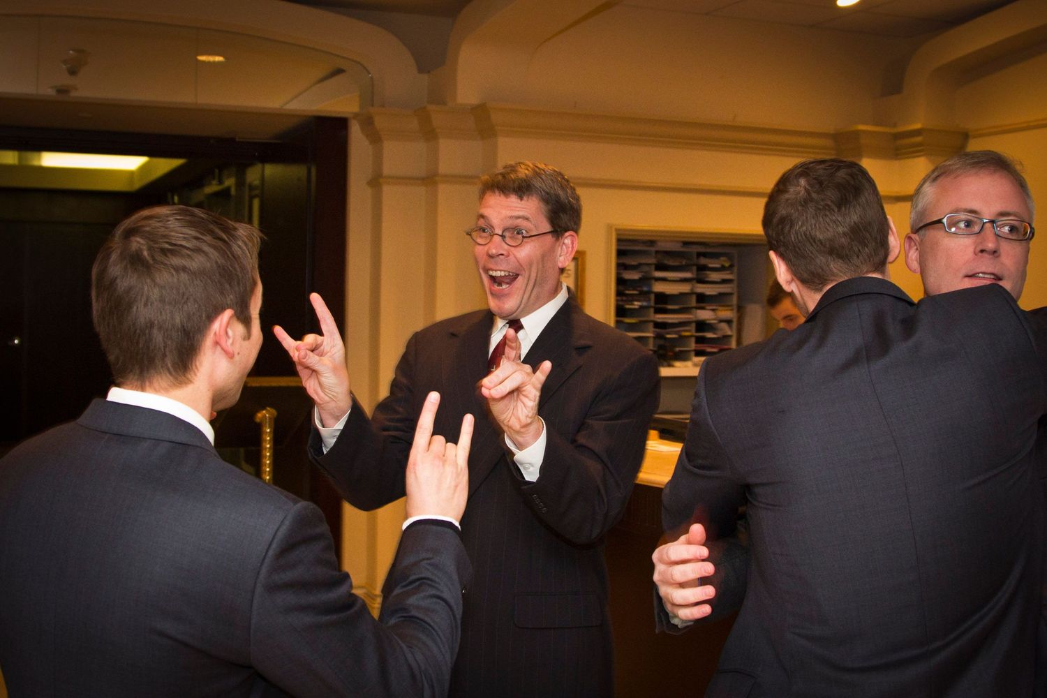 Three men in dark suits laughing and making expressive gestures at a wedding reception.