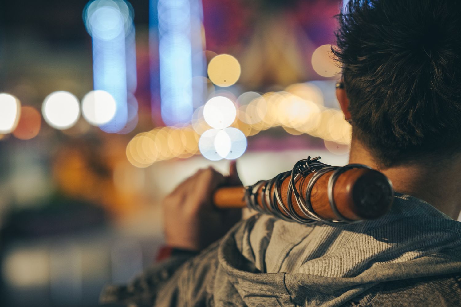 Man seen from behind playing a wooden flute against a blurred night background of glowing golden and blue city bokeh lights.
