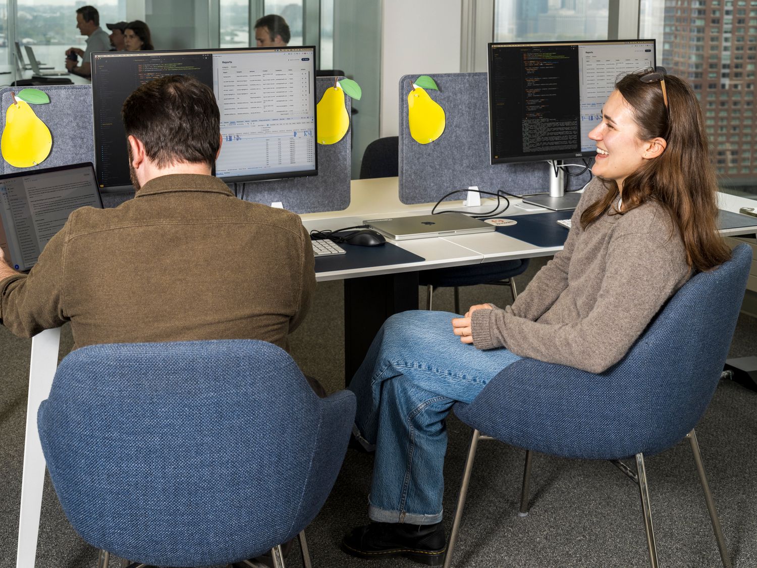 Two office workers sit at desks with monitors displaying a pear logo in a modern open-plan workspace.