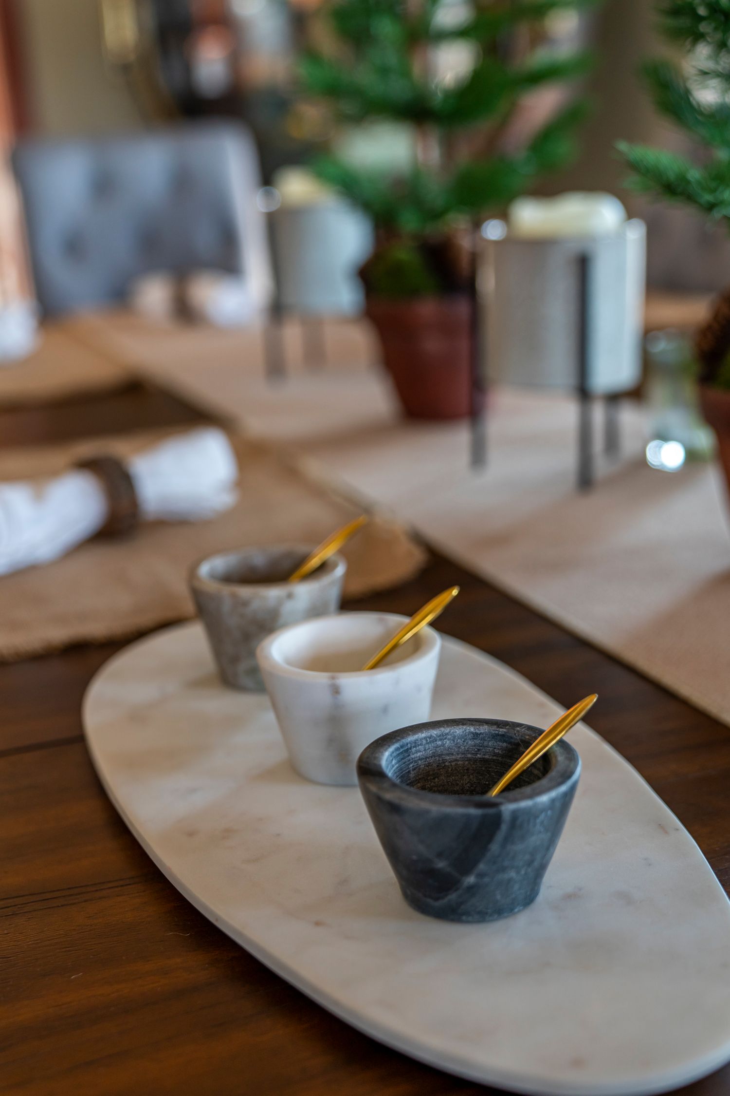 Three marble bowls with gold spoons on an oval marble tray set on a wooden dining table.