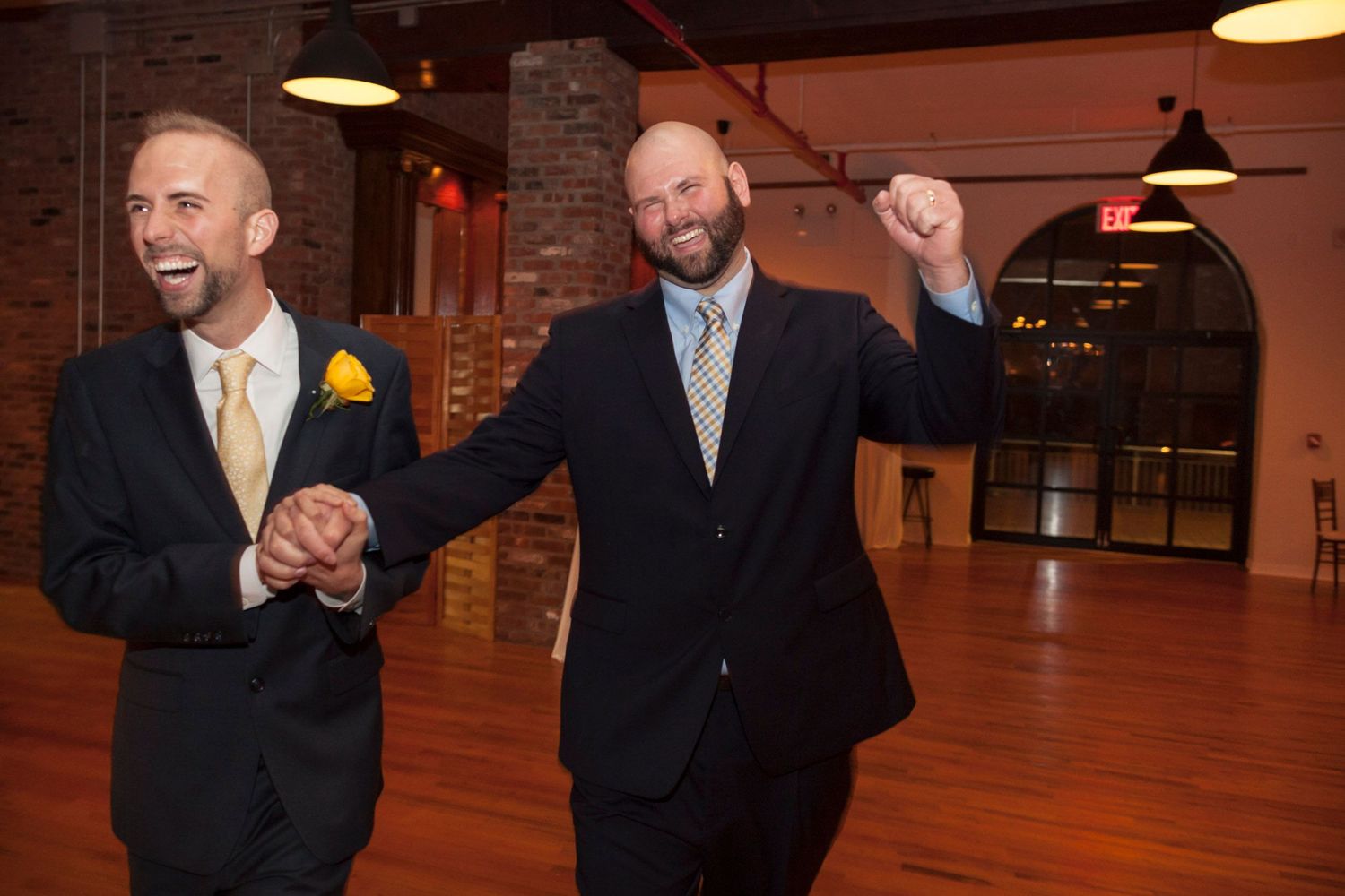 Two grooms in suits celebrate joyfully holding hands at their wedding reception in a brick venue.