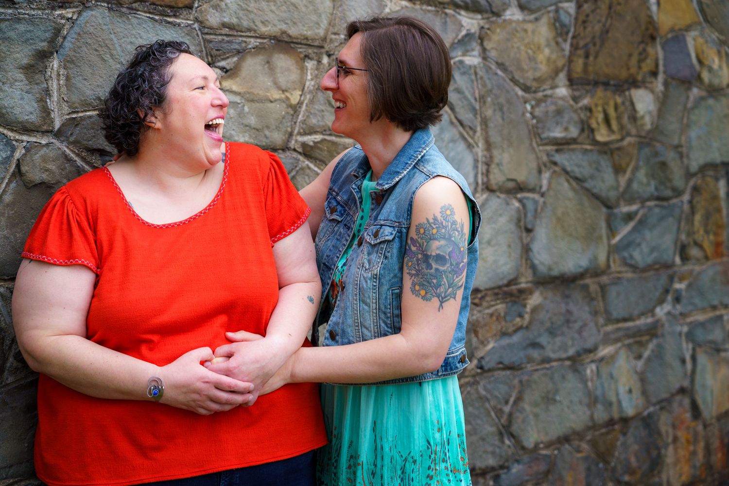 Two women laughing and embracing against a stone wall, one in red top, other in teal skirt with tattoos.