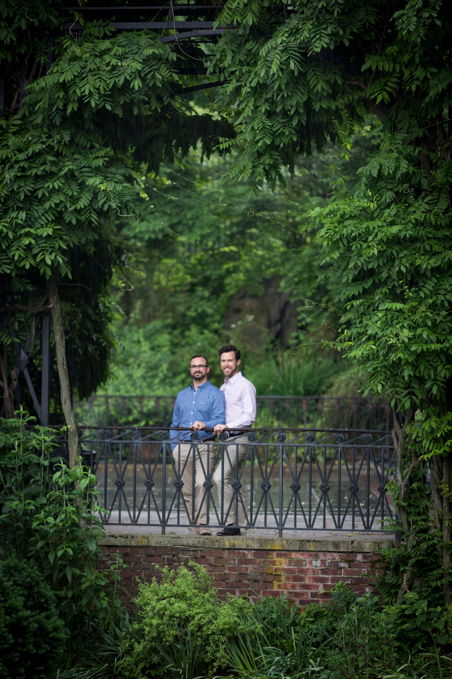 Two men standing on ornate iron bridge surrounded by lush green trees.