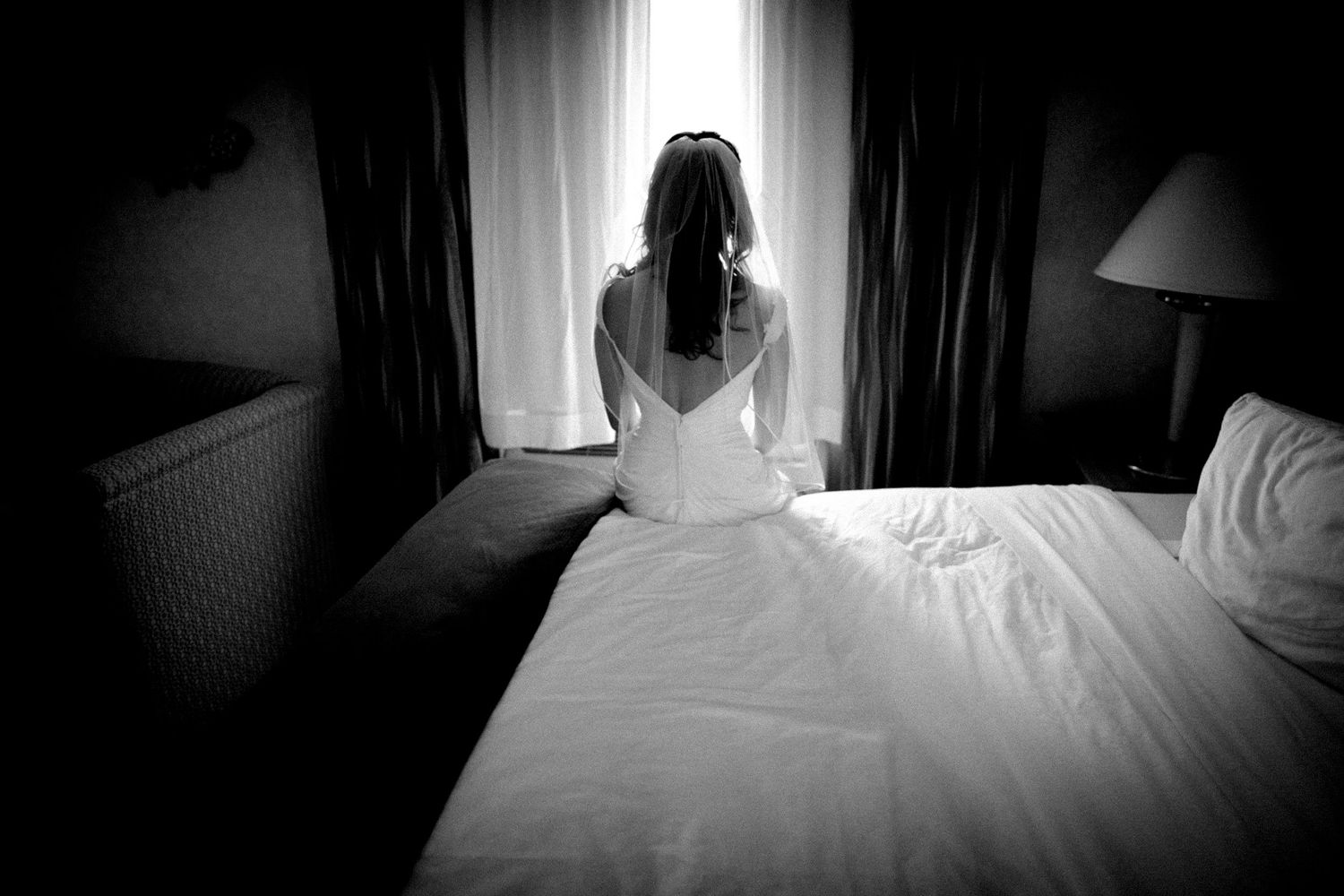 Black and white photo of a bride in wedding dress sitting alone by a hotel room window.