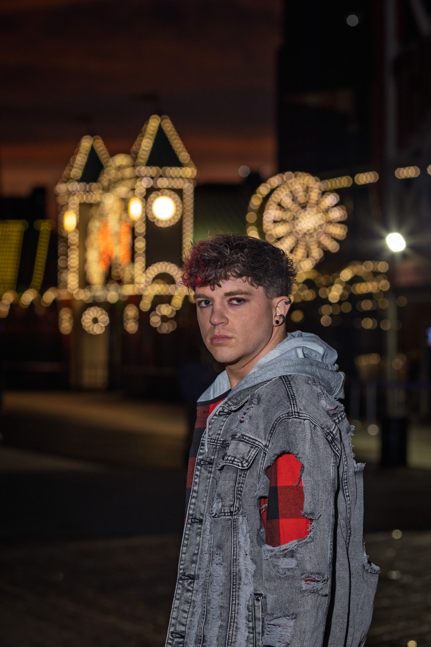 Young man in a distressed gray denim jacket and red plaid shirt poses at night in front of glowing amusement park lights.